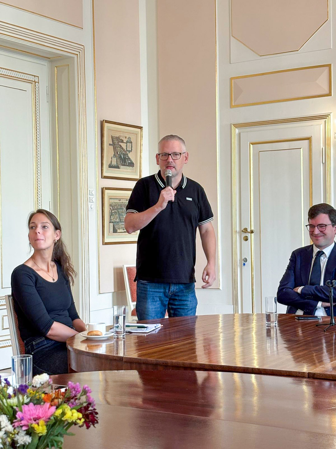 Robert de Louw stoi w rogu pomieszczenia i mówi do mikrofonu. Słuchają go dwie osoby siedzące przy stole