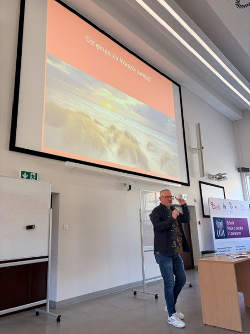 Osoba prowadząca stoi w auli z mikrofonem, kończąc prezentację; na ekranie wyświetlony jest slajd z napisem ‘Dziękuję za Waszą uwagę!’ i zdjęciem morskiego krajobrazu.