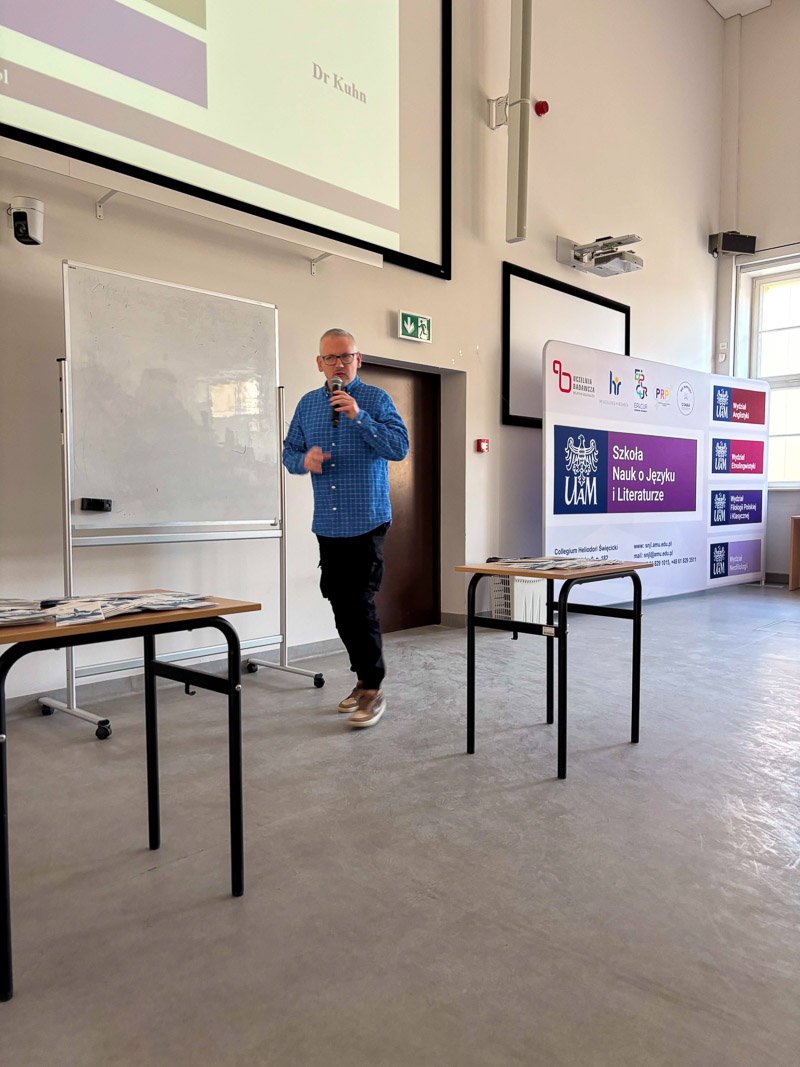 Osoba prowadząca stoi w auli z mikrofonem, mówiąc do publiczności; za nią widoczne są tablice oraz baner Szkoły Nauk o Języku i Literaturze UAM.