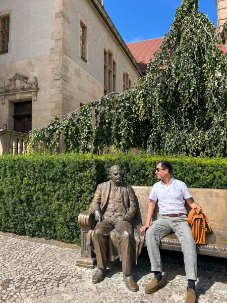 zdjęcie: osoba pozuje do zdjęcia na pamiątkowej ławce na tle gmachu