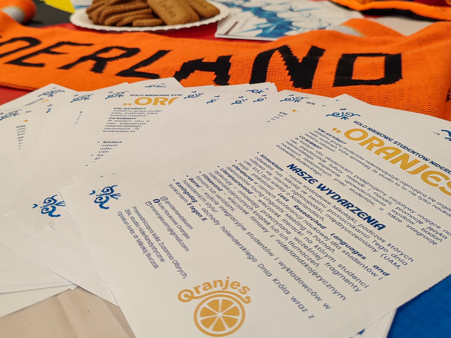 zdjęcie: ulotki Koła Naukowego „Oranjes”, pomarańczowy szalik z napisem „Nederland”, taca z waflami