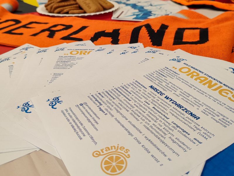 zdjęcie: ulotki Koła Naukowego „Oranjes”, pomarańczowy szalik z napisem „Nederland”, taca z waflami