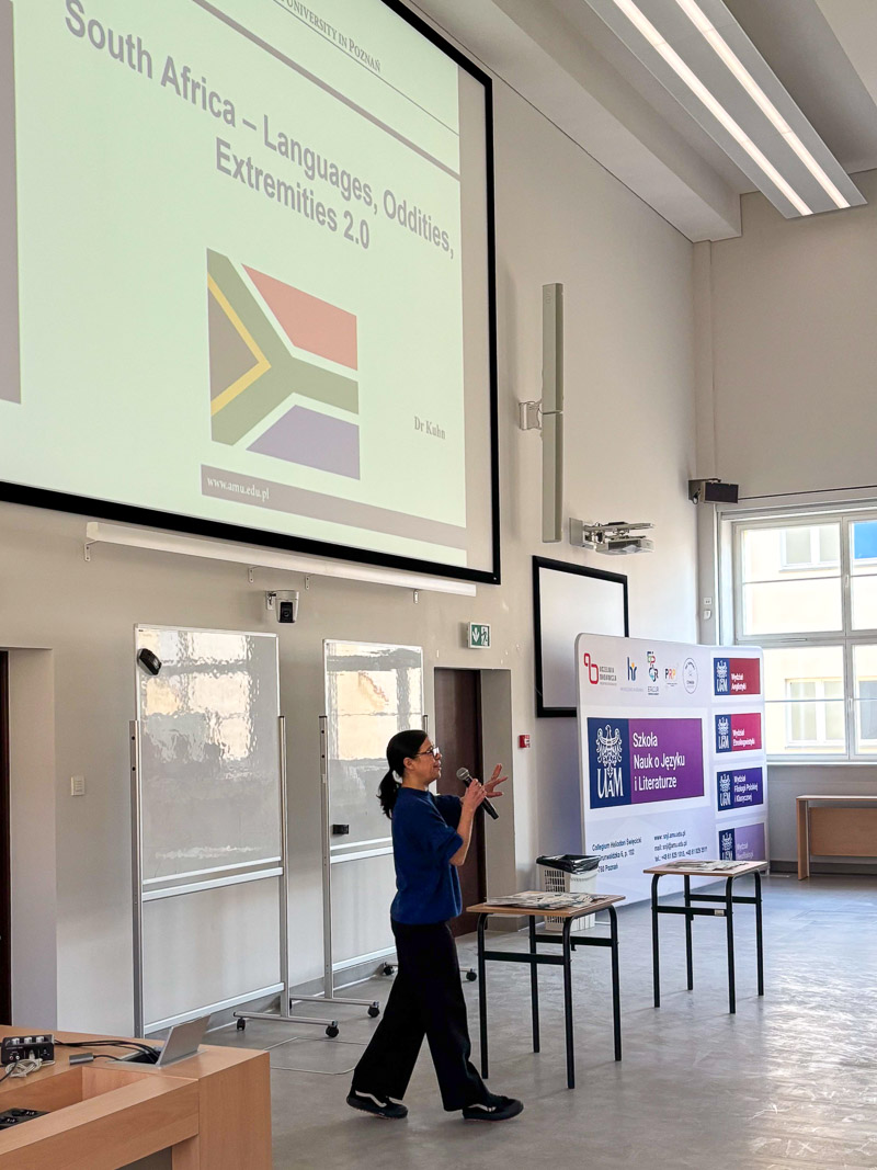 Osoba prowadząca stoi w auli z mikrofonem i omawia slajd zatytułowany ‘South Africa – Languages, Oddities, Extremities 2.0’, wyświetlony na dużym ekranie.