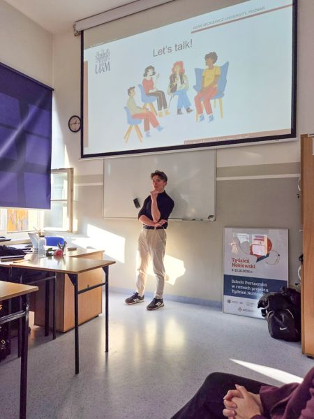 zdjęcie: osoba stoi w sali lekcyjnej; za nią, na ekranie, slajd z prezentacji