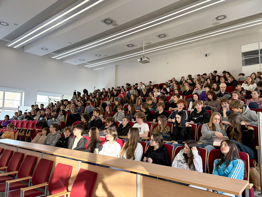 Duża grupa uczestników siedzi w czerwonych fotelach w jasnej auli, oczekując na rozpoczęcie zajęć lub prezentacji.