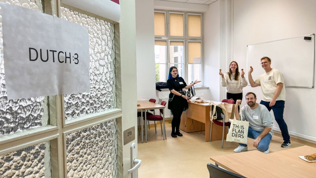 zdjęcie: otwarte drzwi do sali, na których przyklejono kartkę z napisem w języku angielskim {Dutch}, czyli {język niderlandzki}; za drzwiami grupa czworga radosnych osób pozuje przyjaźnie do zdjęcia