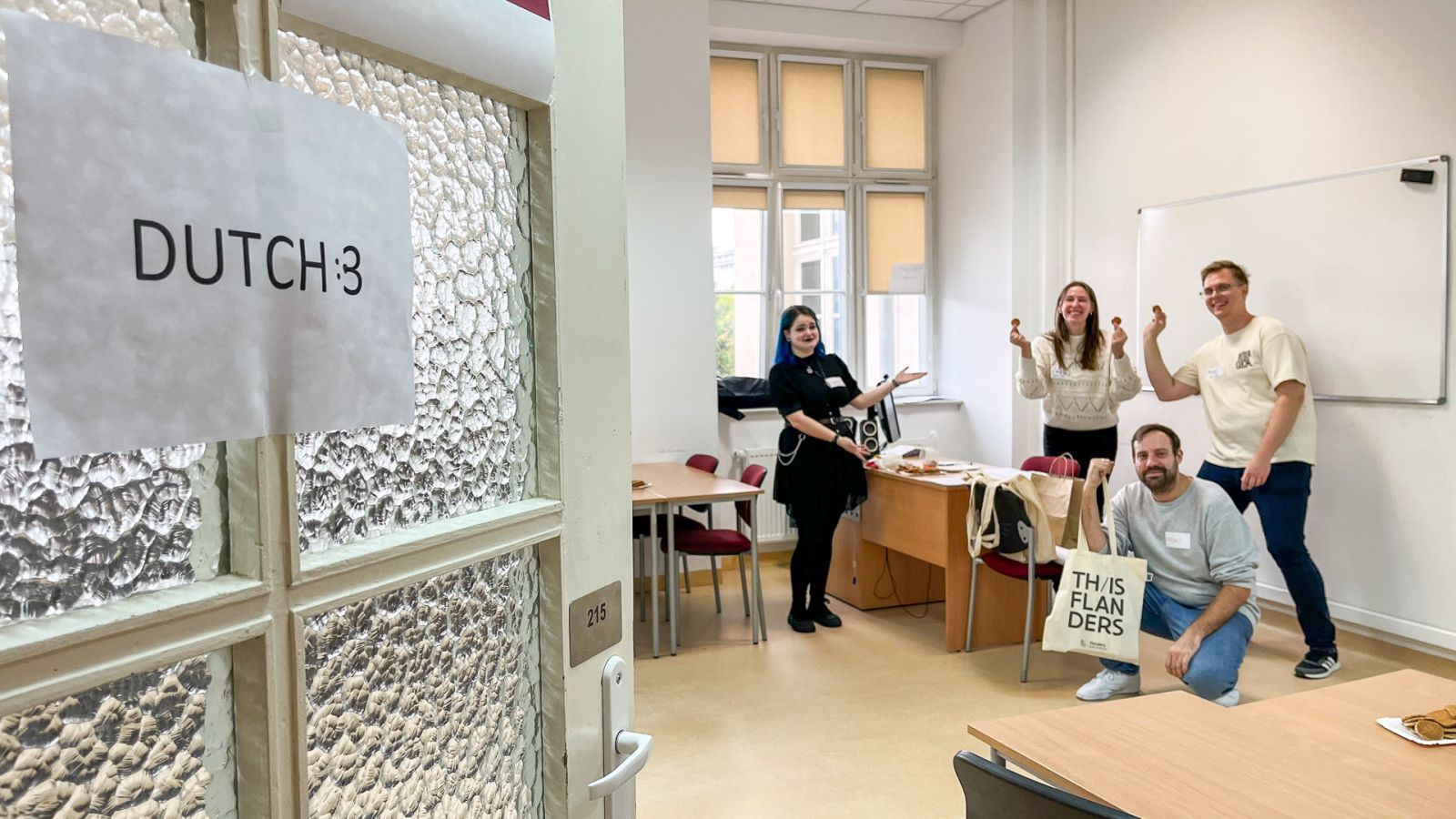 zdjęcie: otwarte drzwi do sali, na których przyklejono kartkę z napisem w języku angielskim {Dutch}, czyli {język niderlandzki}; za drzwiami grupa czworga radosnych osób pozuje przyjaźnie do zdjęcia