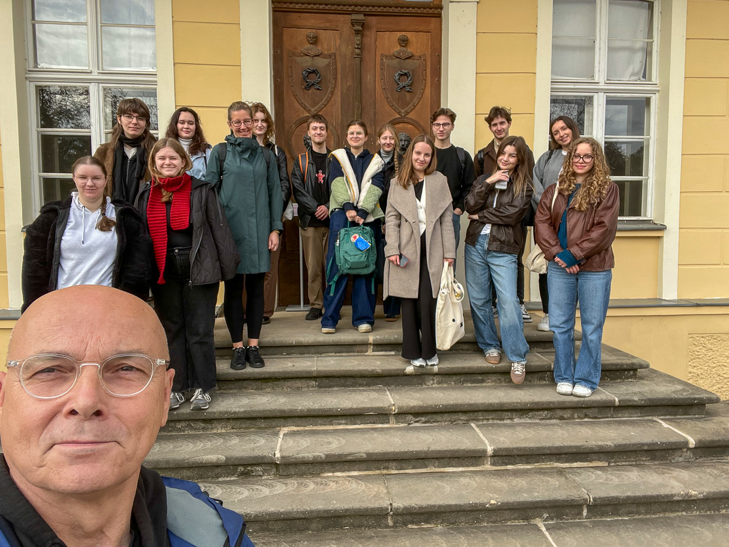 zdjęcie: grupa osób pozuje przed pałacem; jedna osoba jest bliżej kadru, trzyma aparat w ręku