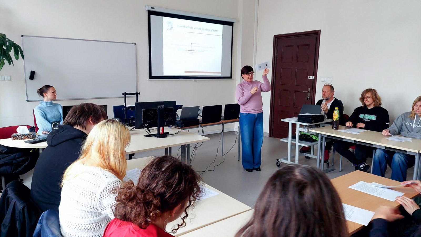 zdjęcie: osoba stoi w sali seminaryjnej; na ekranie za nią: obraz z rzutnika; przed nią: zgromadzona w sali publiczność
