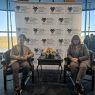 photograph: two people sit in chairs in a large room with a glass facade wall; behind them: a rollup with the logo of the conference