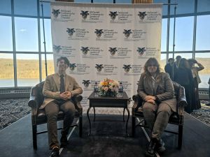 photograph: two people sit in chairs in a large room with a glass facade wall; behind them: a rollup with the logo of the conference