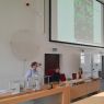 photograph: three persons behind a desk in a lecture hall, two sitting, one standing