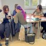 zdjęcie: grupa sześciorga osób siedzi przy stołach w sali zajęciowej, zapisują coś na kawałkach papieru i wymieniają się nimi w ramach warsztatu integracyjnego