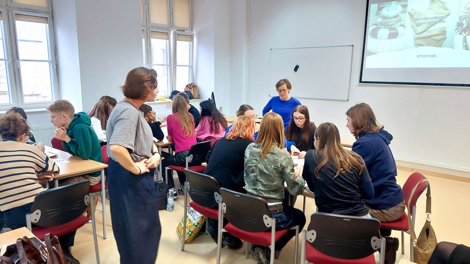 zdjęcie: dwie osoby przemieszczają się po sali seminaryjnej; między nimi: stoły, na których zgromadzone są pomoce dydaktyczne; przy stołach siedzą osoby biorące udział w warsztacie