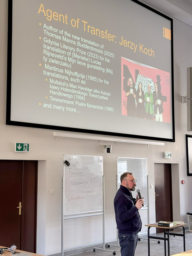 Osoba prowadząca stoi w auli z mikrofonem i omawia slajd dotyczący dorobku Jerzego Kocha, wyświetlony na dużym ekranie.