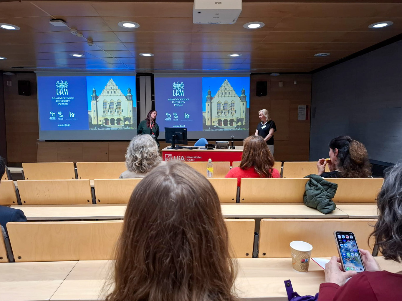 photograph: two persons stand in a lecture hall; behind them: an image projected on a screen from a beamer; in front of them: the audience