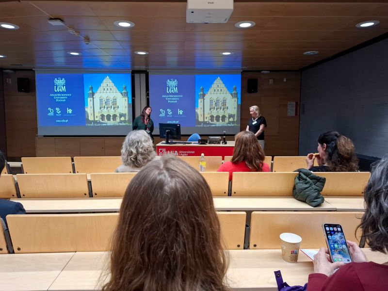 photograph: two persons stand in a lecture hall; behind them: an image projected on a screen from a beamer; in front of them: the audience