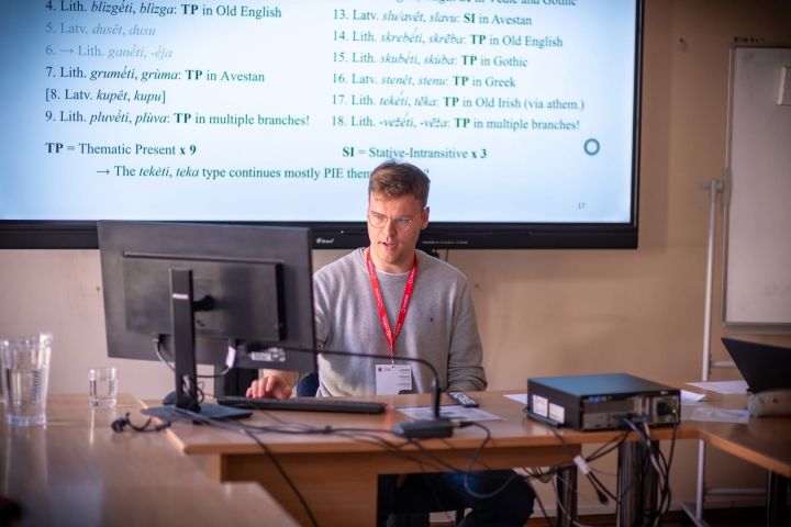 photograph: Sander van Hes behind a desk in a conference room; behind him: a page from a presentation displayed on a screen