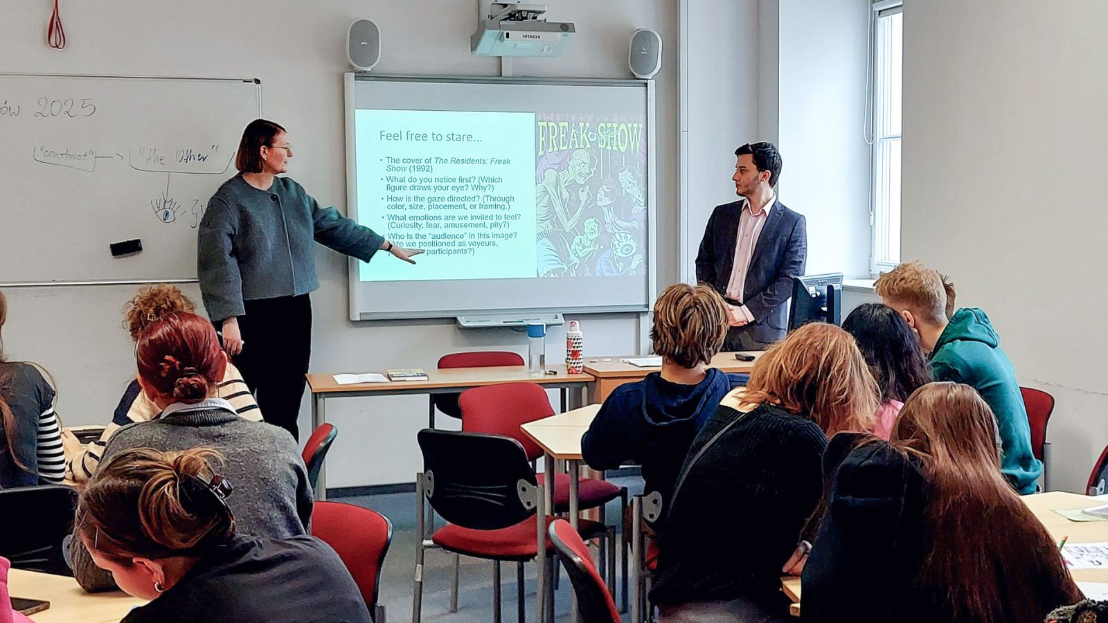 zdjęcie: dwie osoby stoją w sali seminaryjnej; na ekranie za nimi: obraz z rzutnika; przed nimi: zgromadzona w sali publiczność