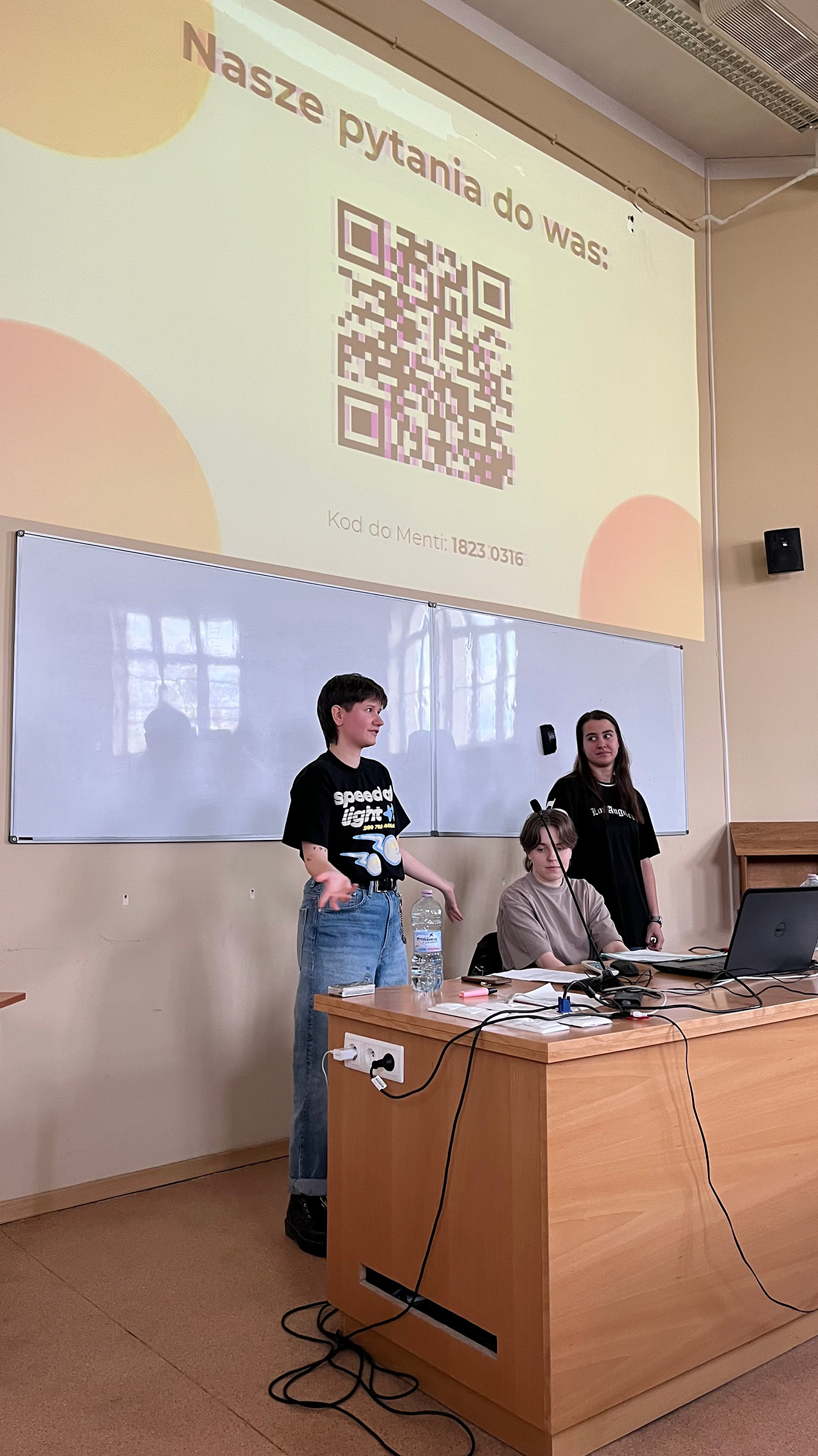 zdjęcie: trzy osoby na tle prezentacji multimedialnej z tekstem „Nasze pytania do Was” i kodem QR