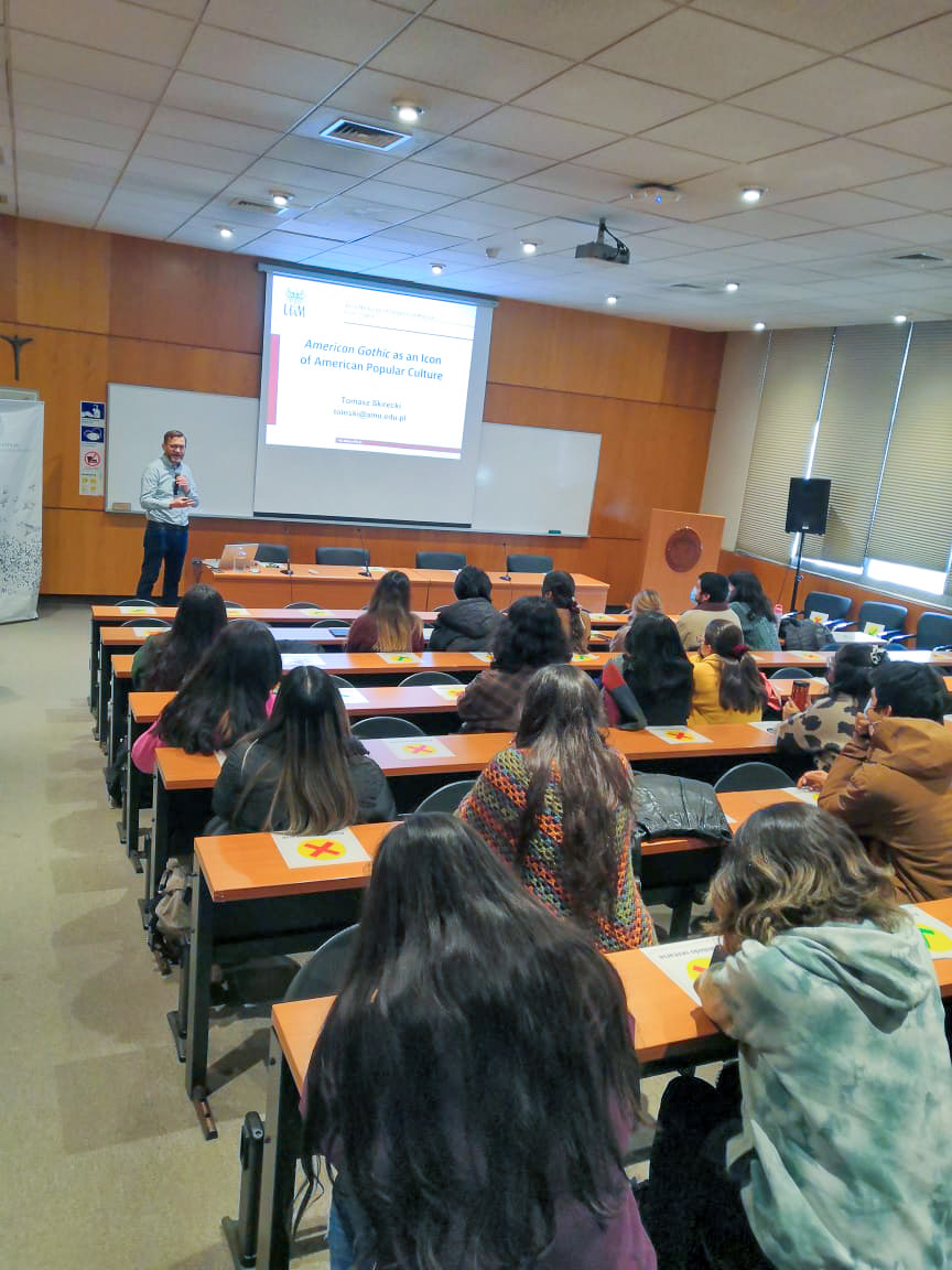 photograph: a person stands in front of an audience in a seminar room; behind them, on a screen: a slide from a slideshow