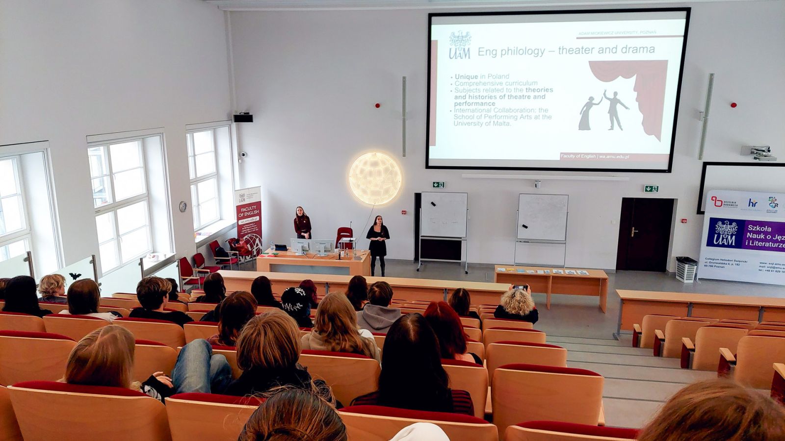 zdjęcie: dwie osoby stoją w sali wykładowej; na ekranie za nimi: obraz z rzutnika; przed nimi: zgromadzona w sali publiczność