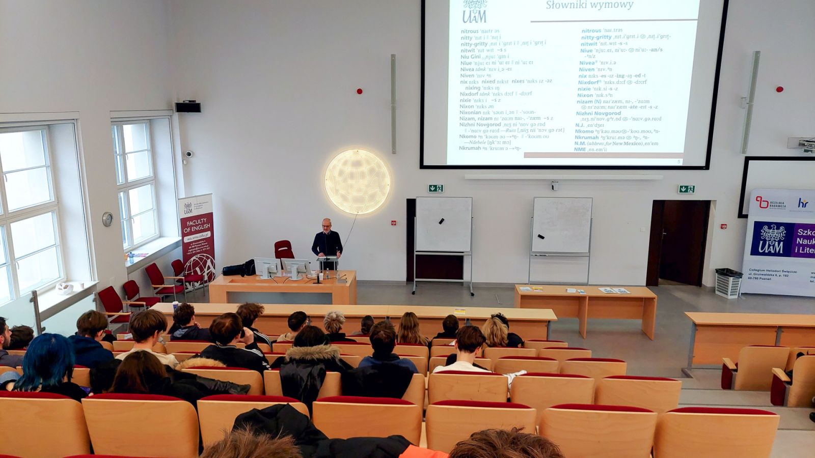 zdjęcie: osoba stoi w sali wykładowej; na ekranie za nią: obraz z rzutnika; przed nią: zgromadzona w sali publiczność