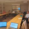 zdjęcie: grupa siedmiu osób siedzi wokół stołu, patrząc w kadr; na pierwszym planie: dwa laptopy, z których jeden wyświetla notatki prelegenta; drugi — slajd