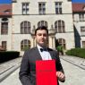 zdjęcie: osoba trzymająca w rękach dyplom pozuje do zdjęcia przed Rektoratem UAM
