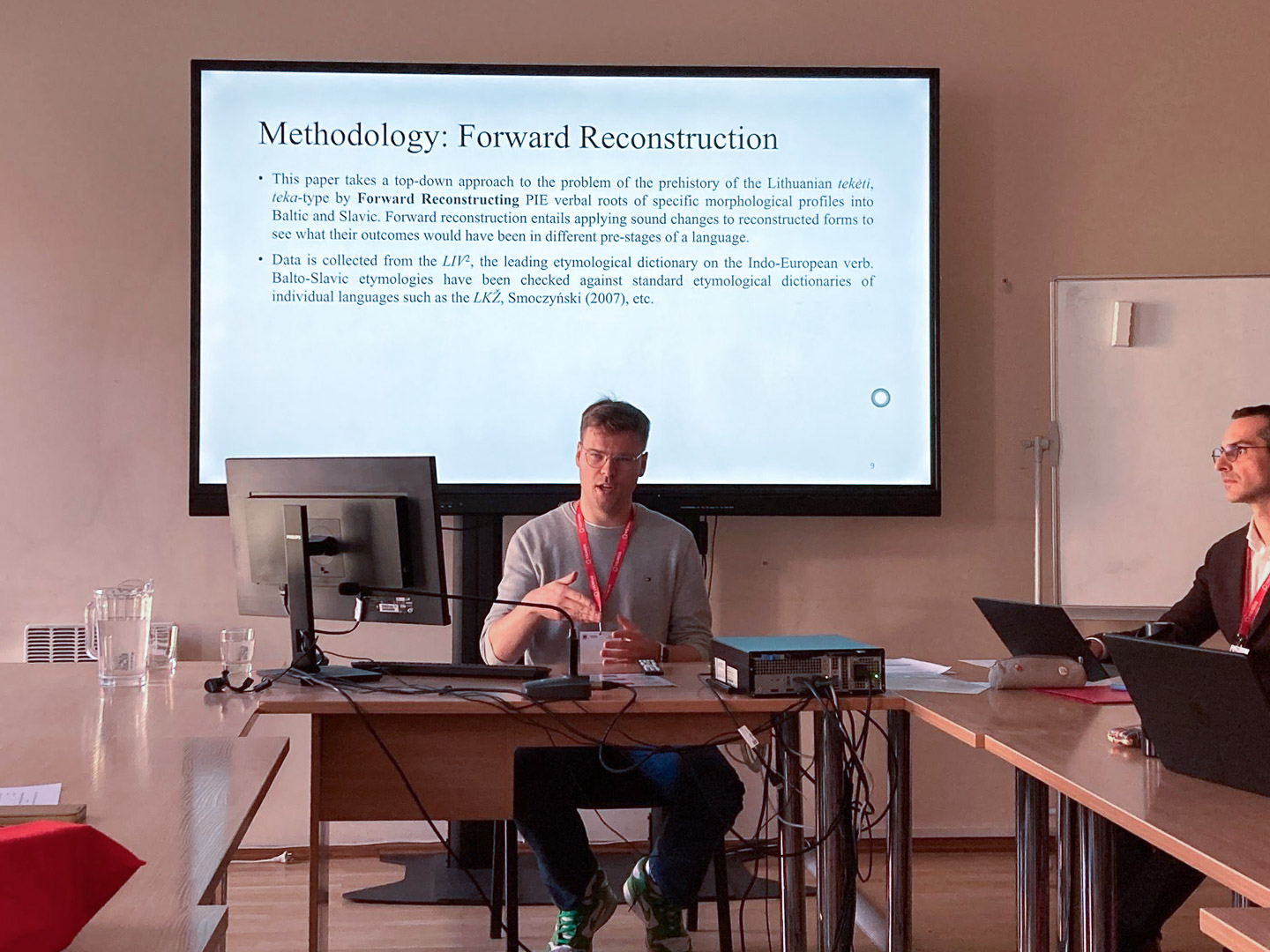 photograph: Sander van Hes behind a desk in a conference room; behind him: a page from a presentation displayed on a screen