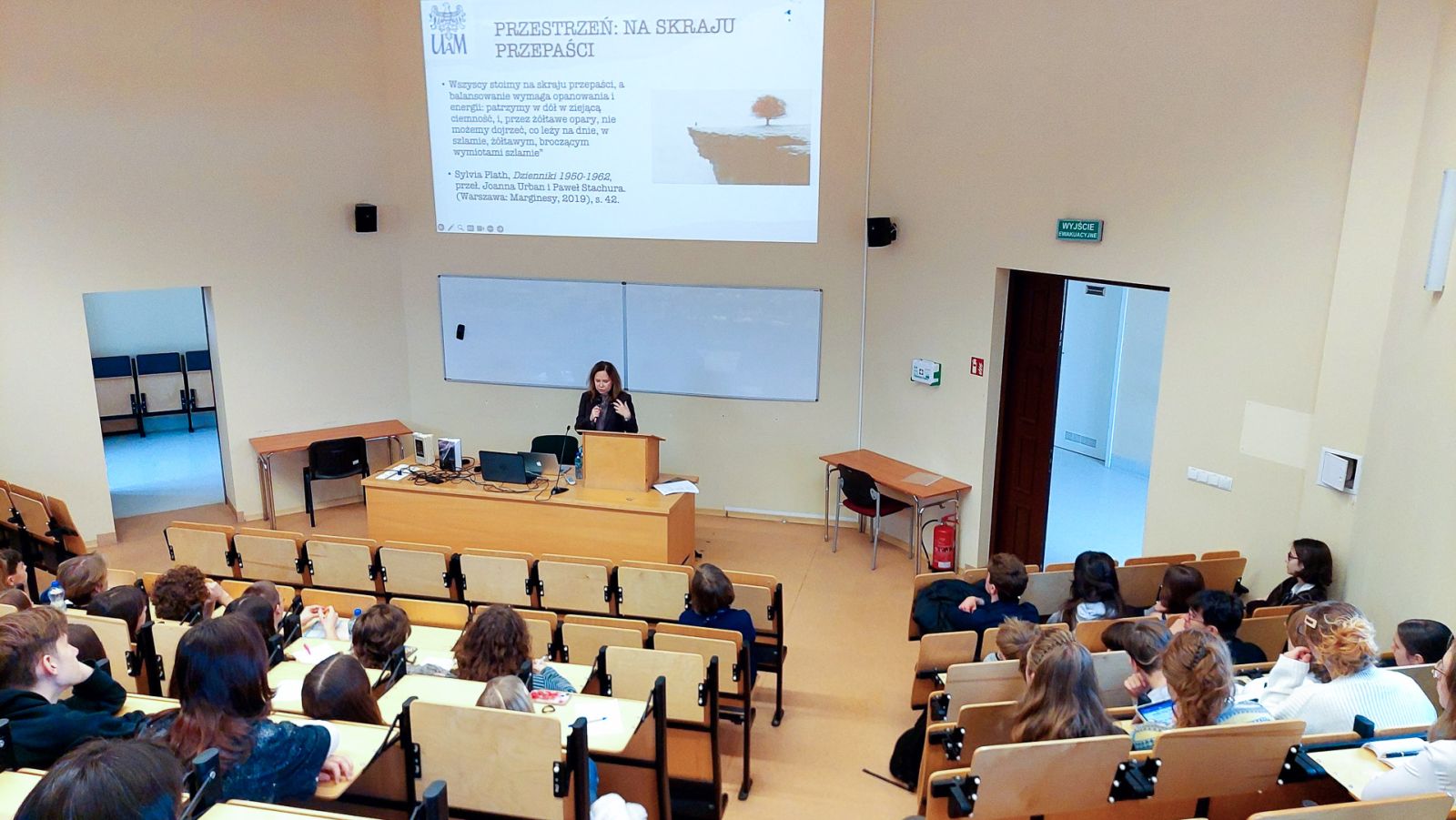 zdjęcie: osoba stoi w sali wykładowej; na ekranie za nią: obraz z rzutnika; przed nią: zgromadzona w sali publiczność