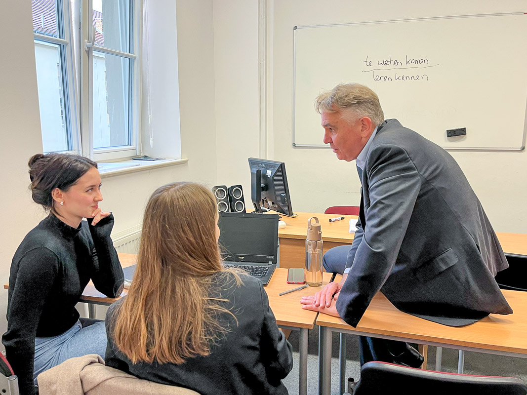 zdjęcie: sala zajęciowa: dwie osoby siedzą za biurkiem, trzecia osoba siedzi na biurku; rozmawiają