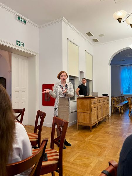 zdjęcie przedstawia postać stojącą w centralnym punkcie sali, gestykulującą, przemawiając do osób zebranych w sali (niewidocznych w kadrze)