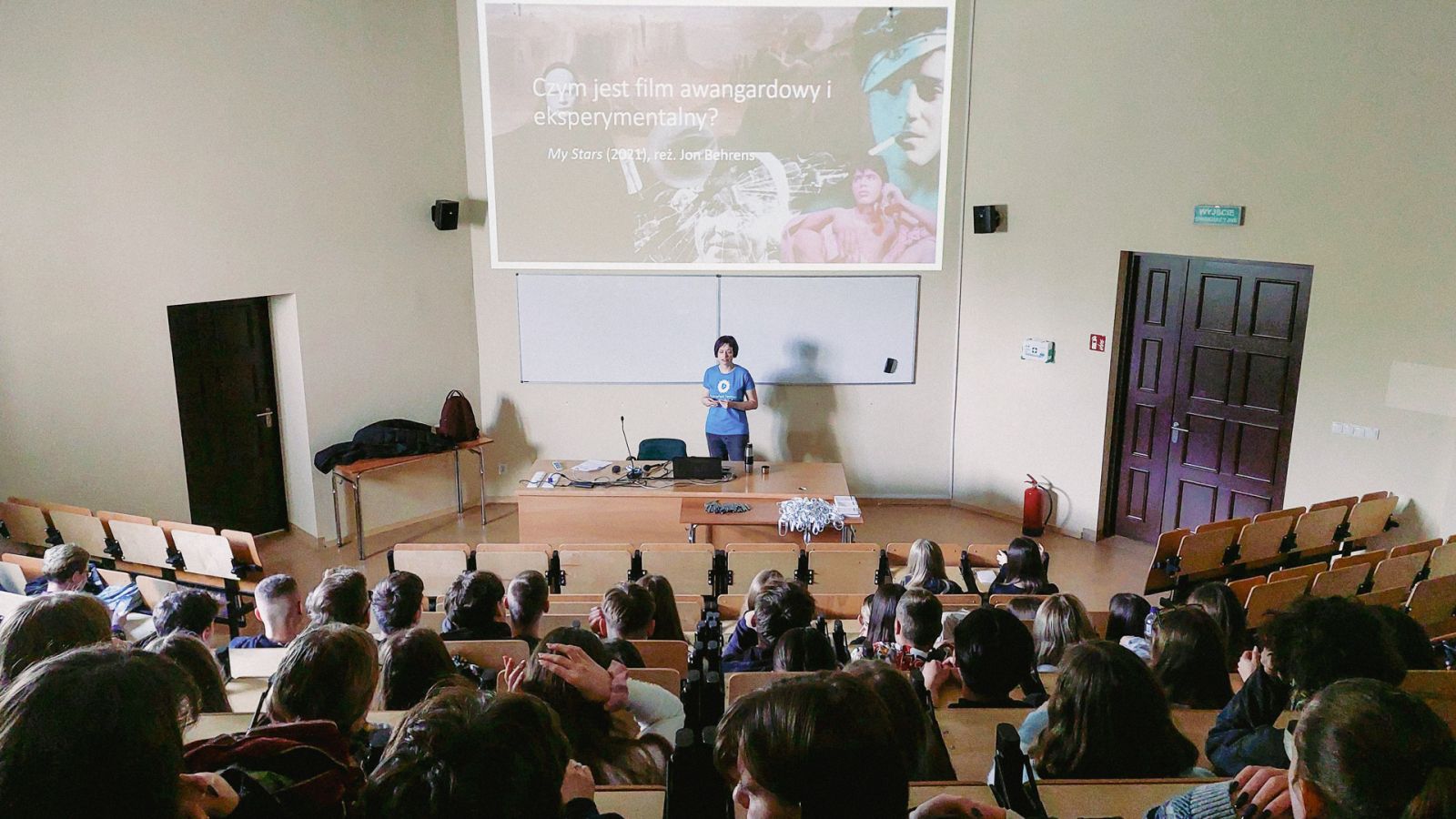 zdjęcie: osoba stojąca przed mównicą w sali wykładowej, na ścianie za nią slajd z prezentacji, przed nią publiczność siedząca w sali