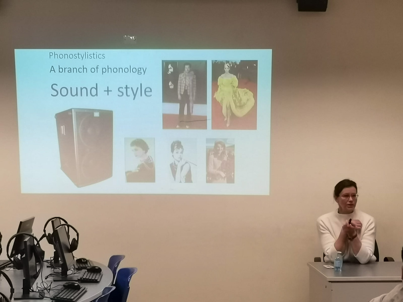 photograph: a person sits in front of an audience in a seminar room (not visible in the image); behind them, on a screen: a slide from a slideshow