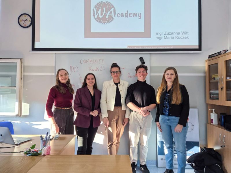 zdjęcie: grupa pięciorga osób stoi w sali lekcyjnej; za nimi, na ekranie, slajd z prezentacji