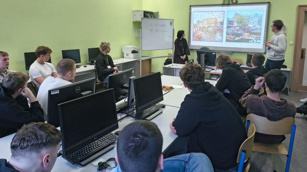 zdjęcie: grupa kilku osób siedzi przy stołach w sali zajęciowej; dwie inne osoby stoją przy tablicy multimedialnej, na której jest wyświetlony slajd z prezentacji