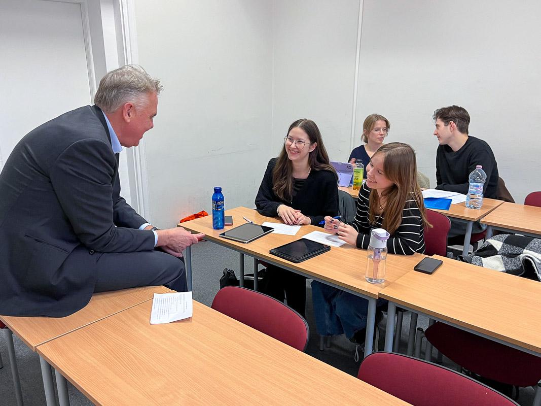 zdjęcie: sala zajęciowa: cztery osoby siedzą za biurkami, piąta osoba siedzi na jednym z biurek; rozmawiają