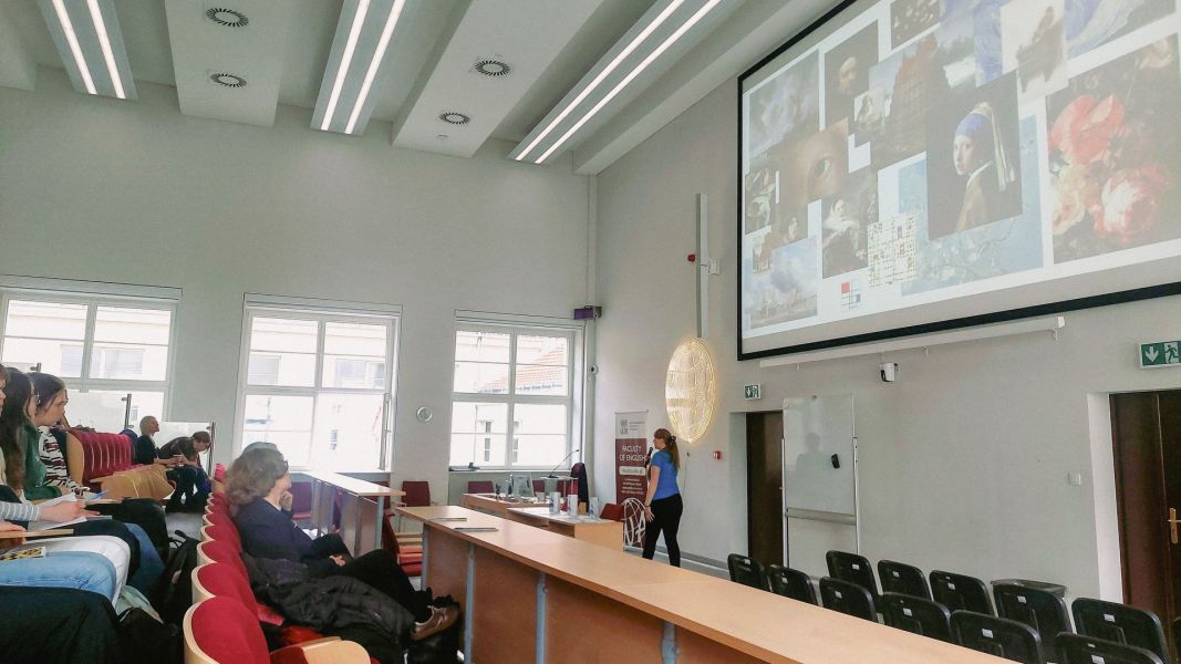 zdjęcie: osoba stojąca przed ekranem w sali wykładowej, na ekranie za nią slajd z prezentacji, przed nią publiczność siedząca w sali