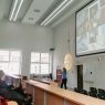 zdjęcie: osoba stojąca przed ekranem w sali wykładowej, na ekranie za nią slajd z prezentacji, przed nią publiczność siedząca w sali