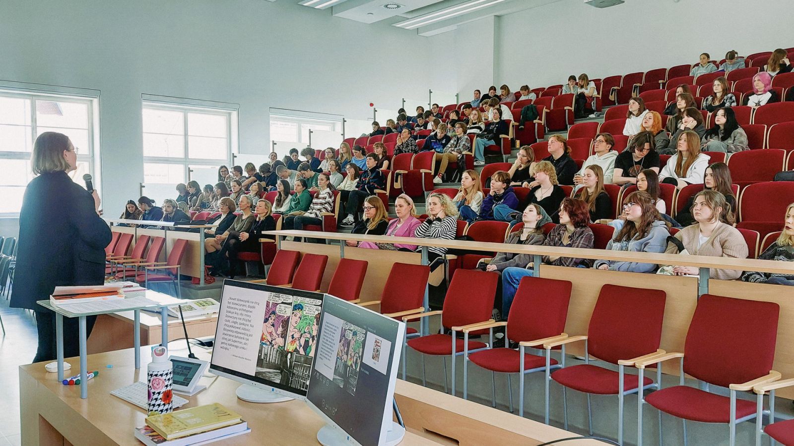 zdjęcie: osoba stojąca za mównicą w sali wykładowej, przed nią publiczność siedząca w sali