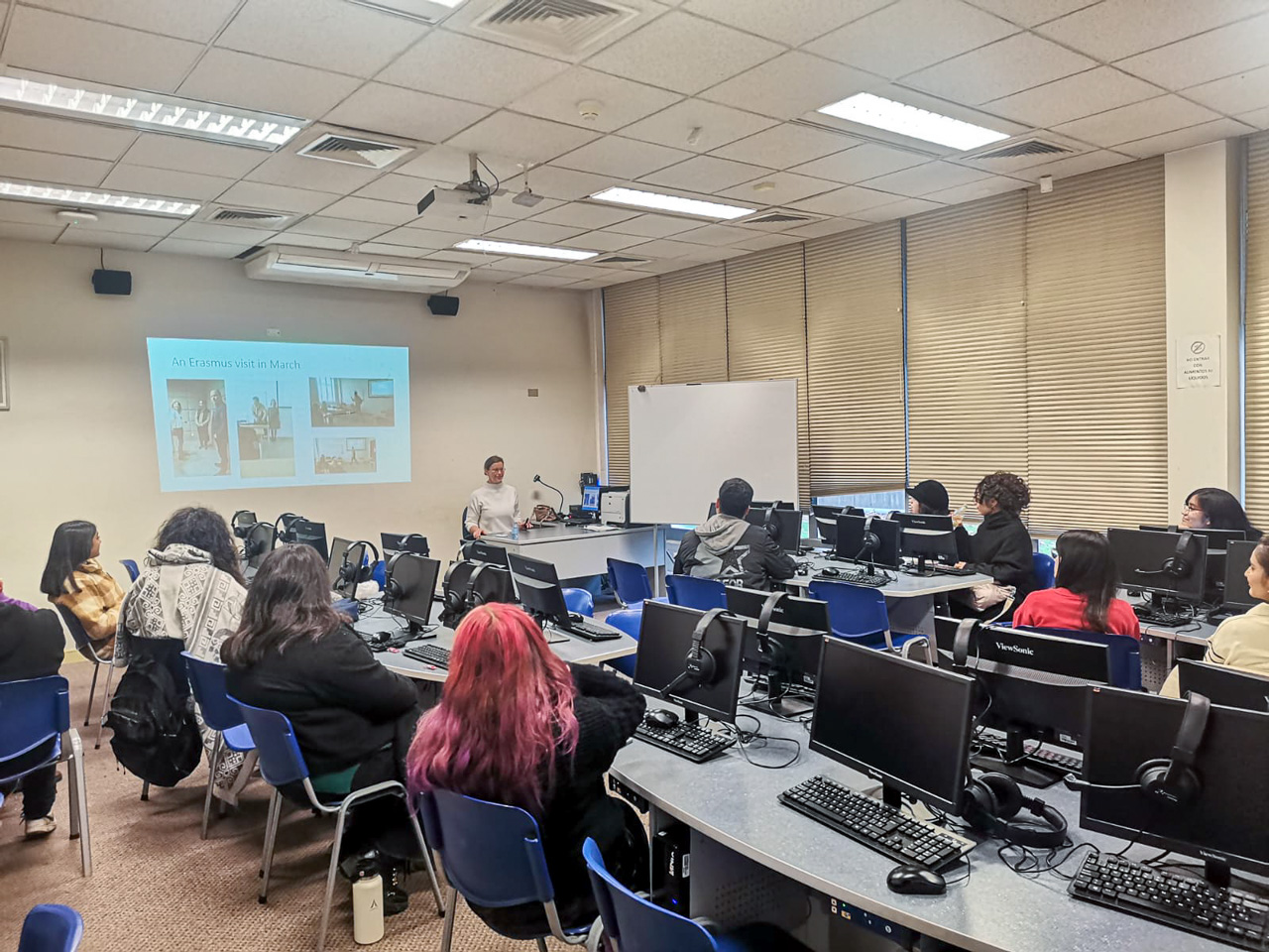 photograph: a person sits in front of an audience in a seminar room; behind them, on a screen: a slide from a slideshow