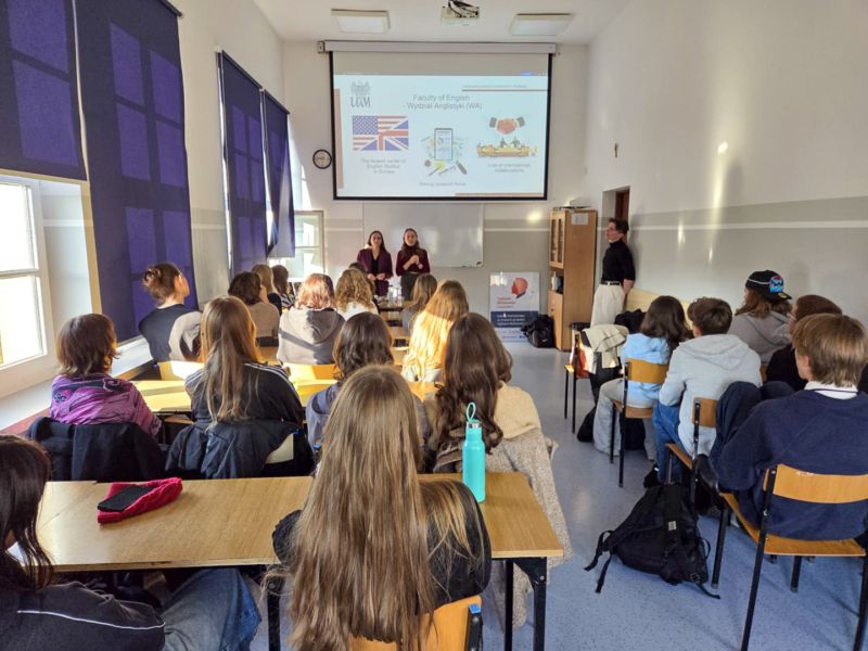 zdjęcie: dwie osoby stoją w sali lekcyjnej, trzecia stoi z boku i przygląda się im; za nimi, na ekranie, slajd z prezentacji; przed nimi: kilkanaście osób siedzi przy ławkach