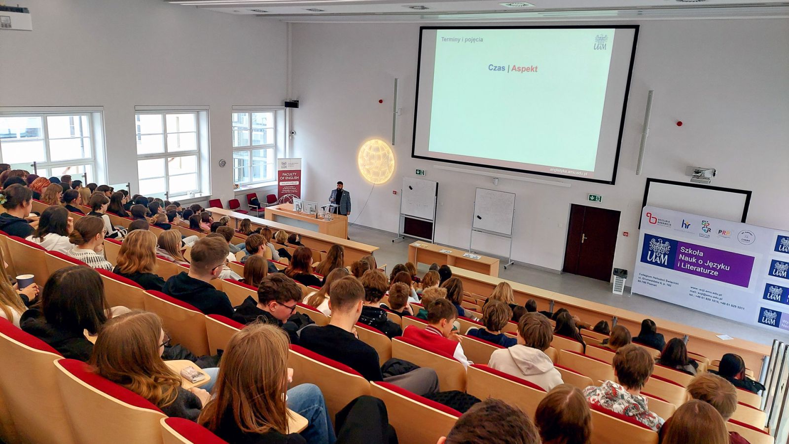 zdjęcie: osoba stoi w sali wykładowej; na ekranie za nią: obraz z rzutnika; przed nią: zgromadzona w sali publiczność