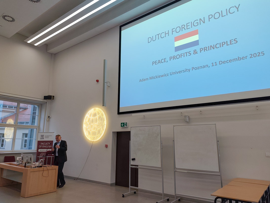 zdjęcie: osoba stoi za mównicą w sali wykładowej; na ścianie za nią: ekran ze slajdem z prezentacji mutlimedialnej