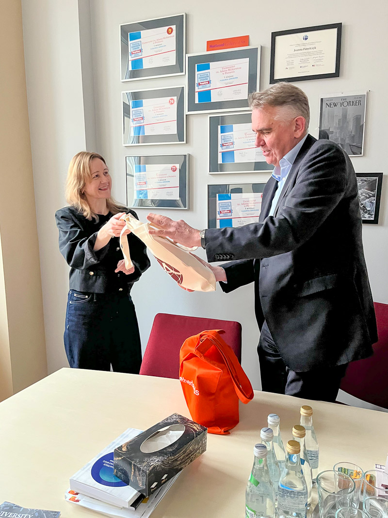 zdjęcie: dwie osoby w pomieszczeniu biurowym wymieniają się okolicznościowymi torbami materiałowymi z upominkami