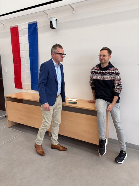 zdjęcie: dwie osoby w auli stoją przy stole, za którym wywieszona jest flaga Królestwa Niderlandów; jedna z osób uśmiecha się w stronę obiektywu