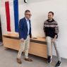 zdjęcie: dwie osoby w auli stoją przy stole, za którym wywieszona jest flaga Królestwa Niderlandów; jedna z osób uśmiecha się w stronę obiektywu