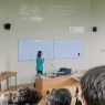 zdjęcie: osoba stojąca przed ekranem w sali wykładowej, na ścianie za nią slajd z prezentacji, przed nią publiczność siedząca w sali