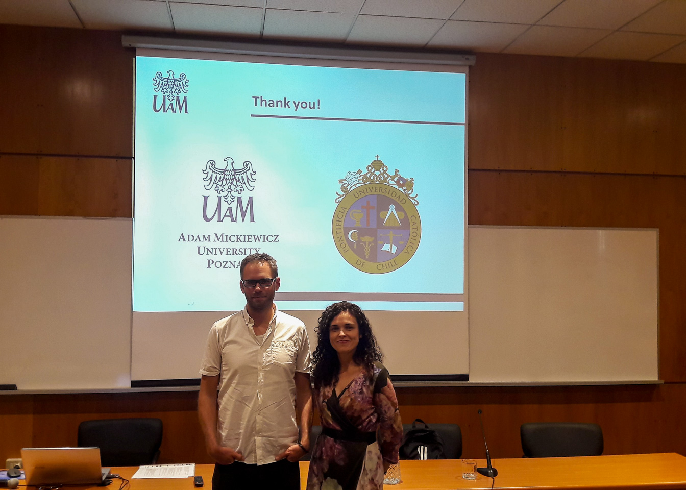 photograph: two persons pose for a photograph; behind them, on a screen: a slide from a slideshow